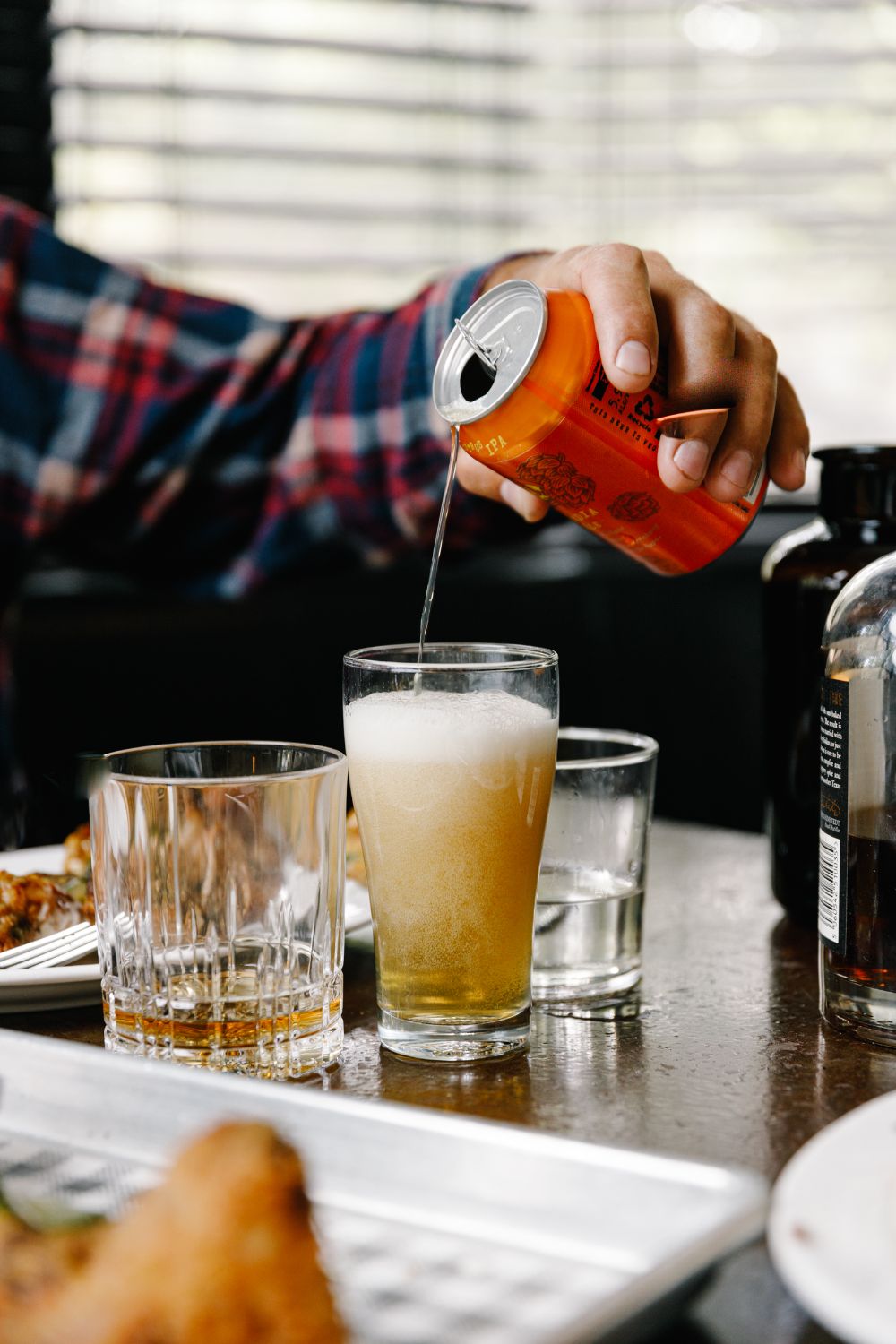 A can of IPA being poured into a glass beside a whisky and water on the table