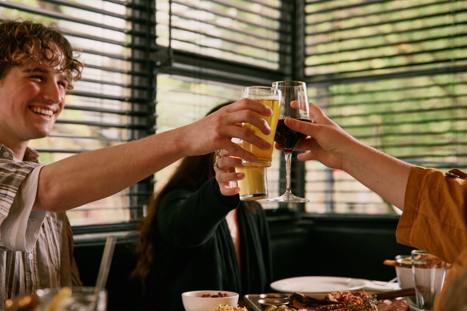 Hands clinking a beer and a glass of red wine over a table of shared BBQ