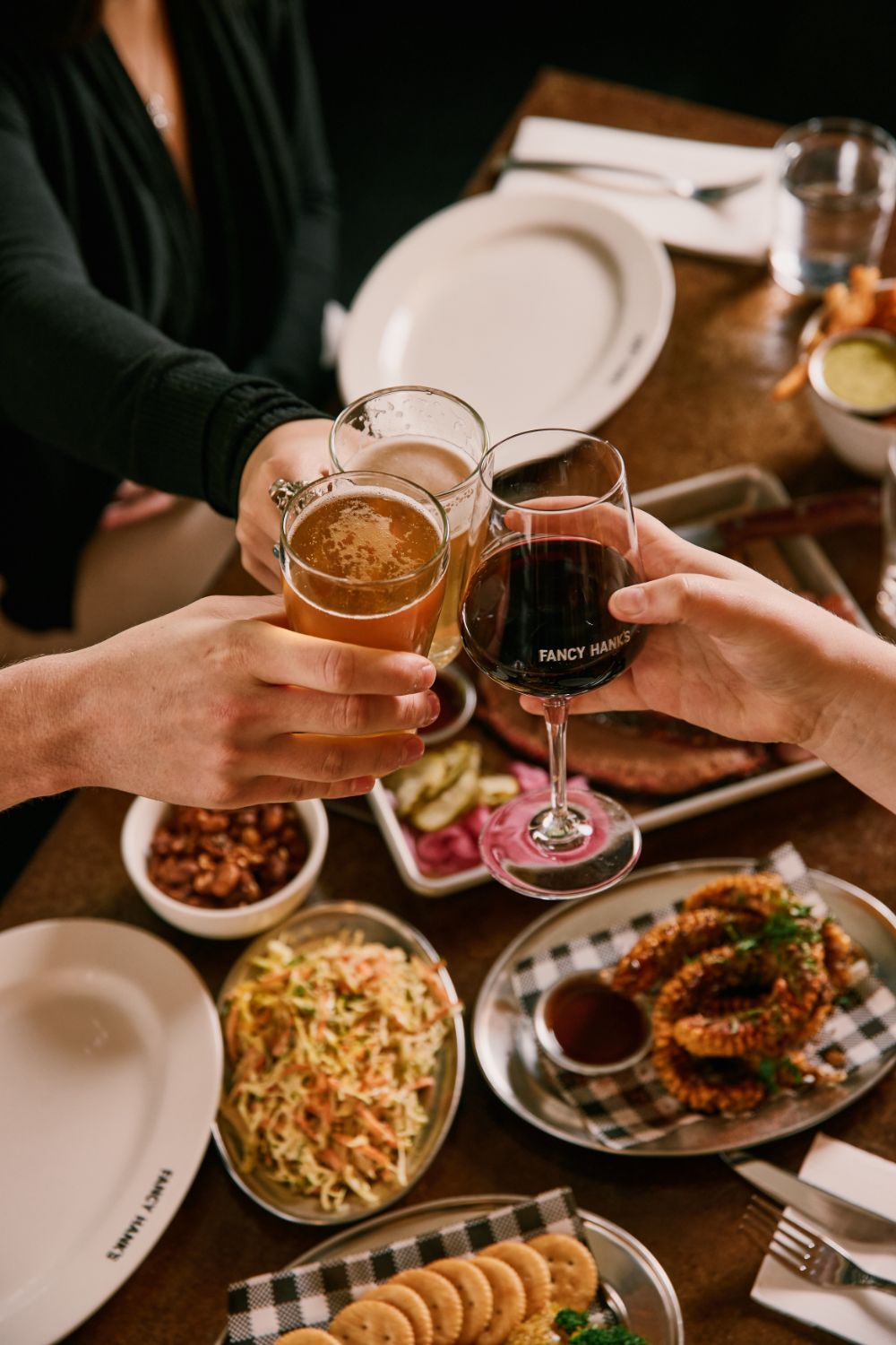 Three friends clinking a beer, a pale ale and a glass of red wine over a table of shared BBQ