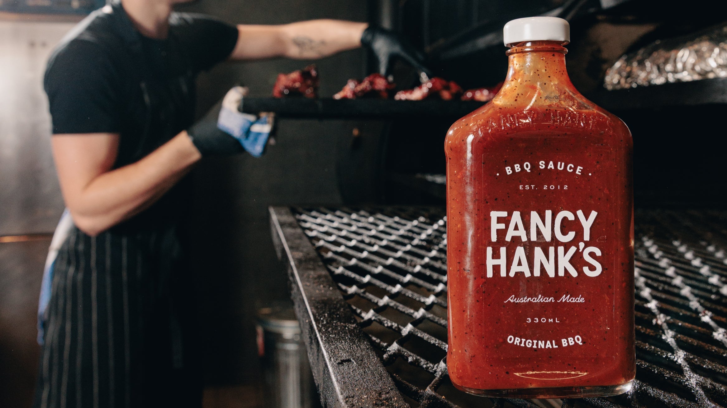 Person grilling with a bottle of Fancy Hank's BBQ Sauce in the foreground.