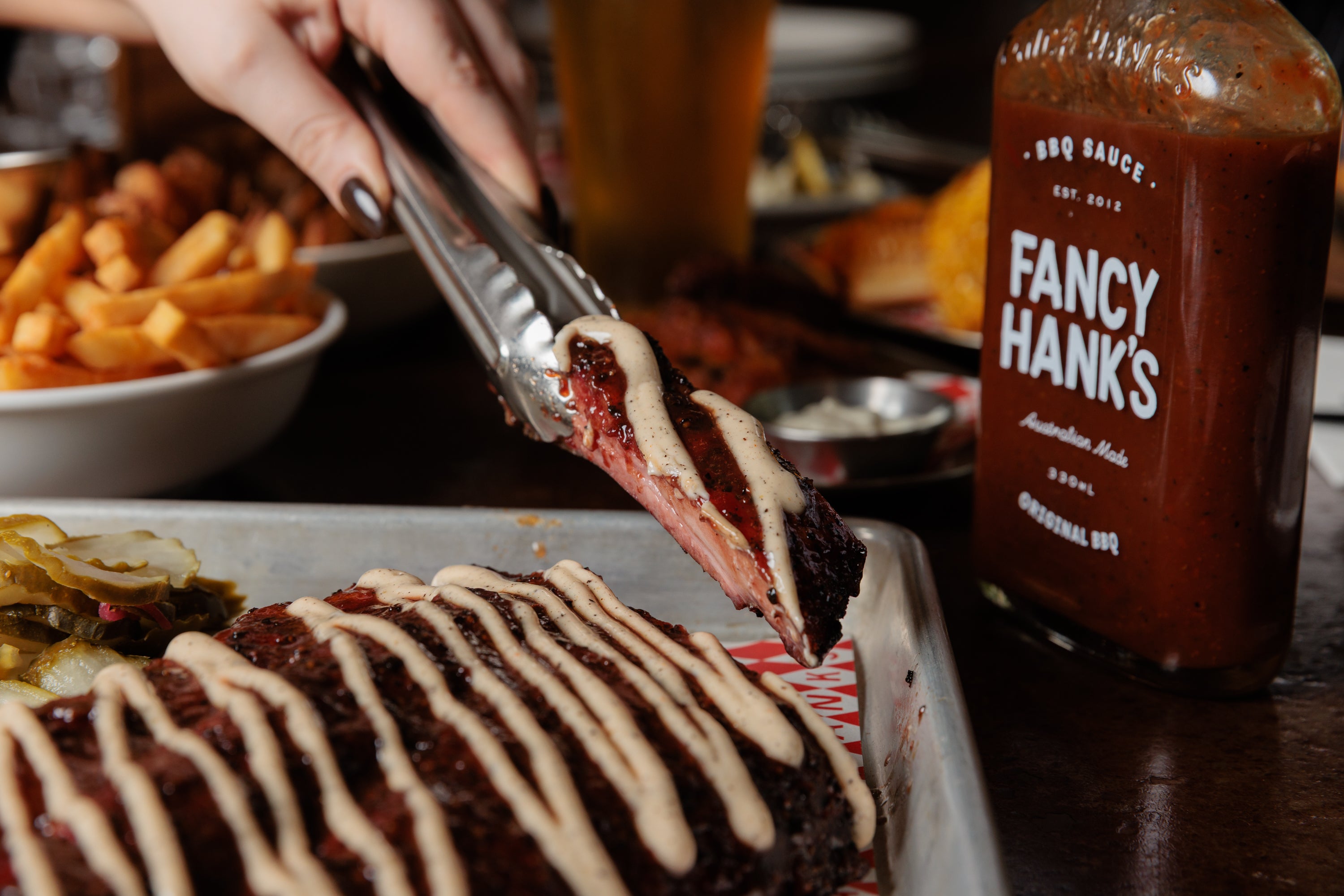 Tongs lifting a sauced pork rib from a tray beside a bottle of Fancy Hank's BBQ sauce