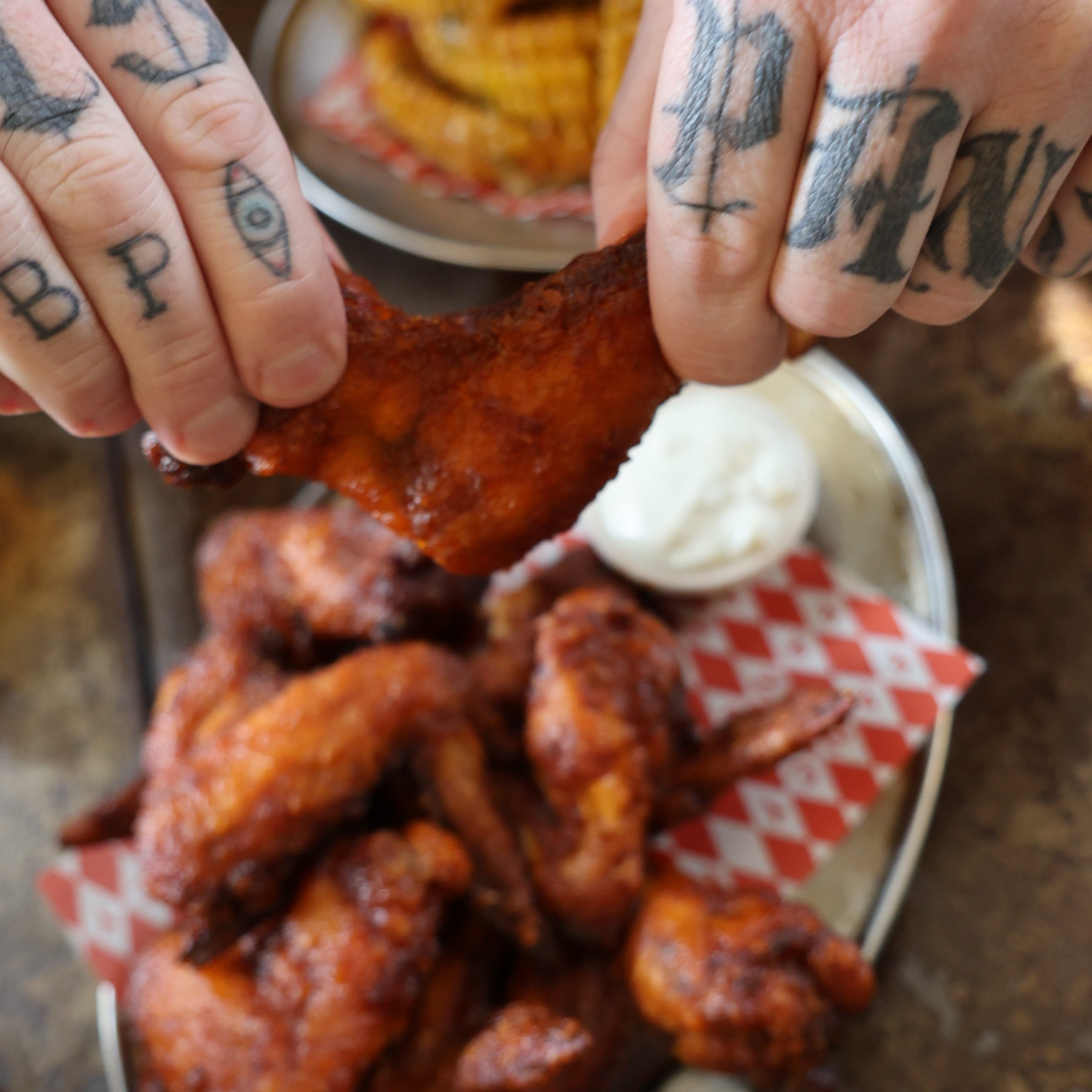 Tattooed hands pulling apart a smoked chicken wing over a plate of wings