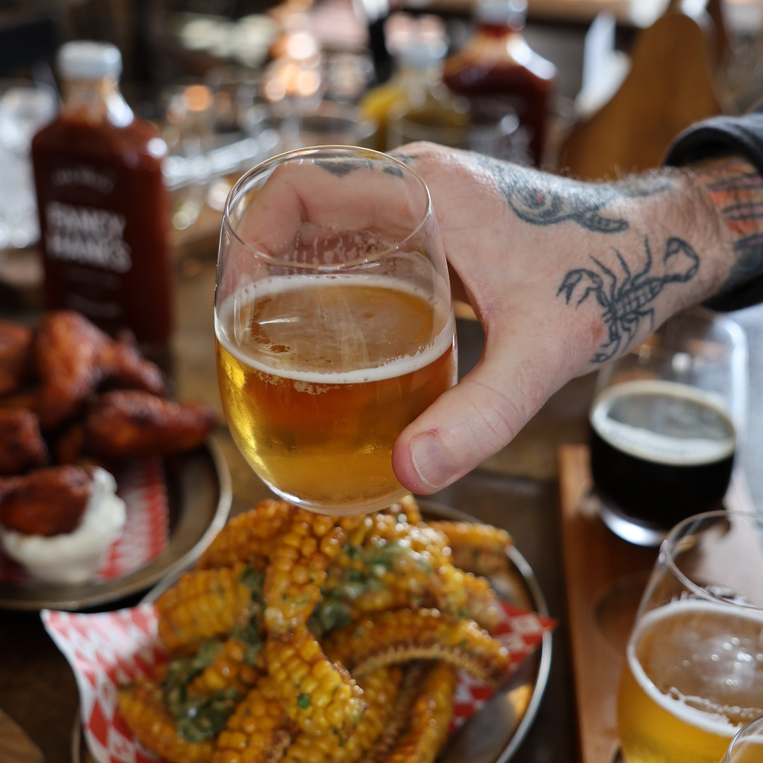 A cold beer lifted over a table of wings and corn ribs